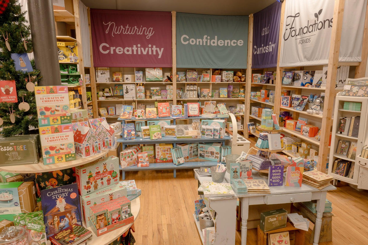 A children's book and toy display inside a bookstore or gift shop, with wooden shelves filled with books, colorful hanging banners reading 'Nurturing Creativity', 'Confidence', and 'curiosity', and a small white table in the foreground with assorted items.